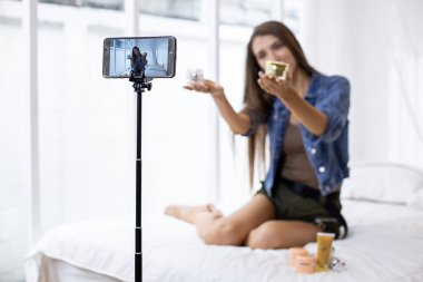 Young woman reviews and selling cosmetics online via her mobile phone on the bed at home. Sell stuff online. Live selling.