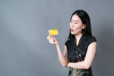Güzel genç Asyalı kadın, gri arka planda elinde kredi kartı olan geleneksel siyah Çin elbisesi giyiyor.