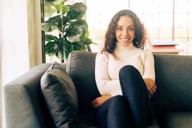 Latin kadın gülümsüyor ve evdeki kanepede oturuyor.