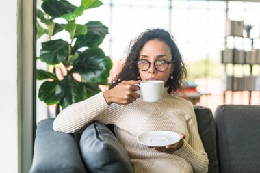 Latin bir kadın evdeki kanepede kahve içiyor. Eski moda efekt filtresi.