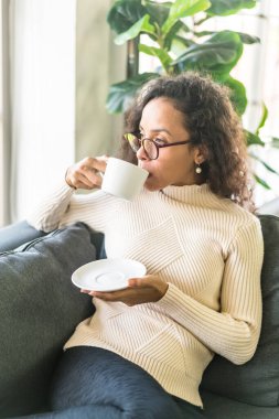 Latin bir kadın evdeki kanepede kahve içiyor. Eski moda efekt filtresi.
