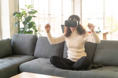 Latin bir kadın evdeki kanepede sanal gerçeklik kulaklığı kullanıyor.