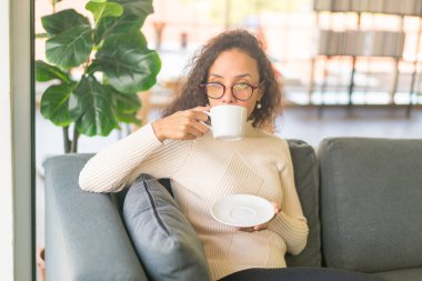 Latin bir kadın evdeki kanepede kahve içiyor. Eski moda efekt filtresi.