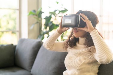 Latin bir kadın evdeki kanepede sanal gerçeklik kulaklığı kullanıyor.