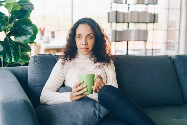 Latin kadın evdeki kanepede kahve içiyor.