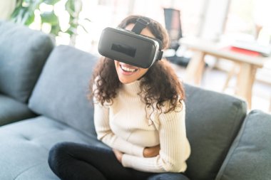 Latin bir kadın evdeki kanepede sanal gerçeklik kulaklığı kullanıyor.