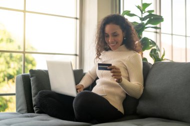 Evdeki kanepede alışveriş yapmak için dizüstü bilgisayar ve kredi kartı kullanan Latin bir kadın.