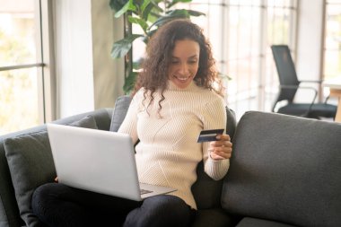 Evdeki kanepede alışveriş yapmak için dizüstü bilgisayar ve kredi kartı kullanan Latin bir kadın.