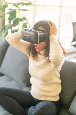 Latin bir kadın evdeki kanepede sanal gerçeklik kulaklığı kullanıyor.
