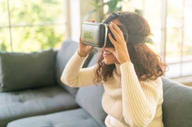 Latin bir kadın evdeki kanepede sanal gerçeklik kulaklığı kullanıyor.