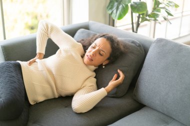 Latin kadın evdeki kanepede sırtı ağrıyan bir şekilde yatıyor.