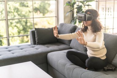 Latin bir kadın evdeki kanepede sanal gerçeklik kulaklığı kullanıyor.