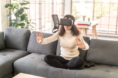 Latin bir kadın evdeki kanepede sanal gerçeklik kulaklığı kullanıyor.