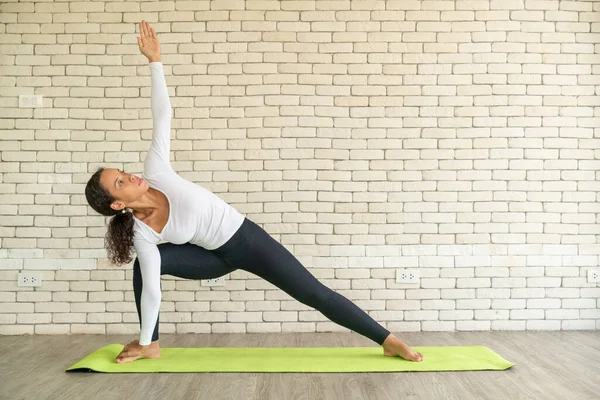Latin woman practicing yoga on mat with white brick background