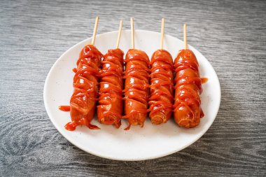 Beyaz tabakta ketçaplı kızarmış sosis şişi.