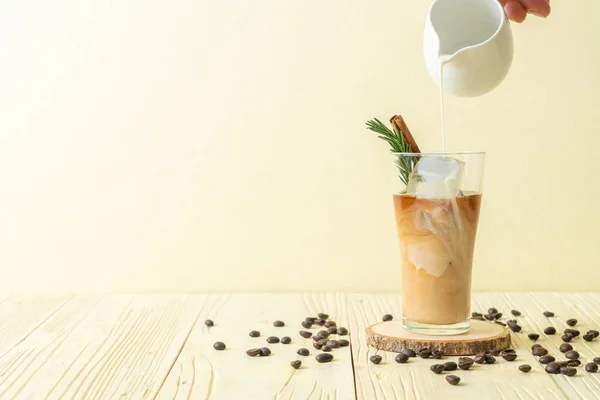 pouring milk in black coffee glass with ice cube, cinnamon and rosemary on wood background