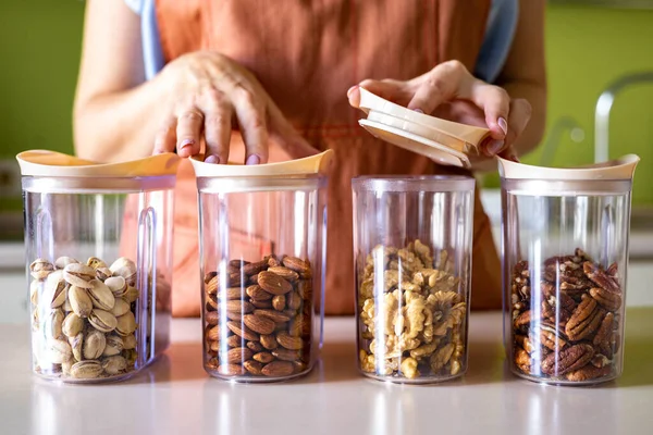 Κοντινό πλάνο γυναίκα χέρια σε ποδιά τοποθέτηση διαφορετικών ξηρών καρπών σε γυάλινο βάζο δοχείο αποθήκευσης στην κουζίνα — Φωτογραφία Αρχείου
