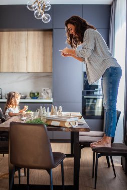 Genç bir kadın sandalyeye çıkıp mutfaktaki Noel kompozisyonunun en iyi görüntülerini çekiyor.