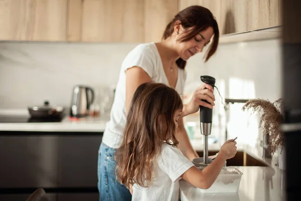 Mamá e hija preparan glaseado para el pan de jengibre en su cocina casera. Batir con una licuadora. La chica ayuda a la mujer. — Foto de Stock