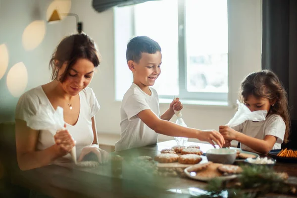 Mom and children decorate Christmas gingerbread at home. A boy and a girl paint with cornets with sugar icing on cookies. New Years decor, branches of a Christmas tree. — Stock Photo, Image