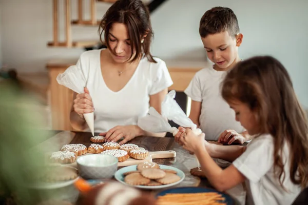 Mamma e bambini decorano il pan di zenzero di Natale a casa. Un ragazzo e una ragazza dipingono con cornetti con la glassa di zucchero sui biscotti. Decorazione di anno nuovo, rami di un albero di Natale. — Foto Stock