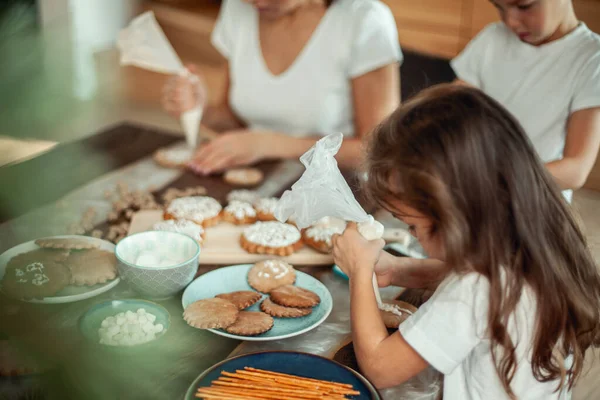 Mamma e bambini decorano il pan di zenzero di Natale a casa. Un ragazzo e una ragazza dipingono con cornetti con la glassa di zucchero sui biscotti. Decorazione di anno nuovo, rami di un albero di Natale. — Foto Stock