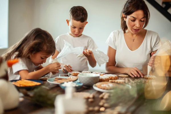 Mom and children decorate Christmas gingerbread at home. A boy and a girl paint with cornets with sugar icing on cookies. New Years decor, branches of a Christmas tree. — Stock Photo, Image