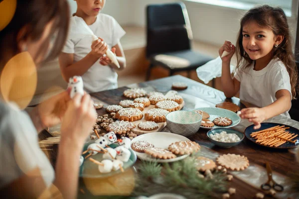 Mamma e bambini decorano il pan di zenzero di Natale a casa. Un ragazzo e una ragazza dipingono con cornetti con la glassa di zucchero sui biscotti. Decorazione di anno nuovo, rami di un albero di Natale. — Foto Stock