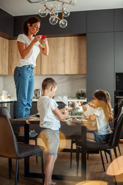 Mom takes pictures of topview as daughter and son decorate gingerbread with sugar icing. Stylish home dining and kitchen. — Stock Photo, Image