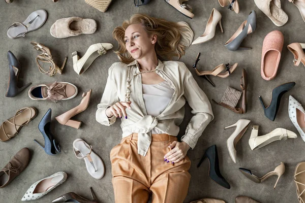 Top view stylish blonde female posing choosing shoes sitting on floor. Top view many female shoes on gray floor fashion stylish footgear choosing