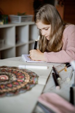 Küçük tatlı bir kız albümde çizim kalemleriyle dairesel bir mandala deseni çiziyor..