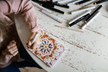 Küçük tatlı bir kız albümde çizim kalemleriyle dairesel bir mandala deseni çiziyor..
