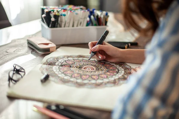 Jovem mulher bonita está sentada na cozinha da casa e desenhando uma mandala para colorir. Desenho zen. — Fotografia de Stock