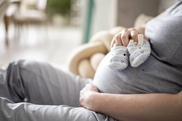 Κοντινό πλάνο έγκυος γυναίκα κρατώντας χαριτωμένα παιδικά μποτάκια στην τεράστια κοιλιά χαλαρώνοντας στο σπίτι περιμένοντας το μωρό — Φωτογραφία Αρχείου