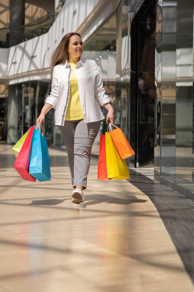 Happy young shopaholic plus size female going at shopping mall holding multicolored paper bags