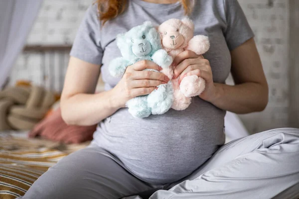 Szczęśliwy ciężarna kobieta siedzi na łóżku w sypialni gospodarstwa dwa słodkie niedźwiedź zabawki czeka bliźniaki dziecko — Zdjęcie stockowe