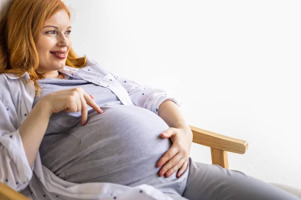 Κοντινό πλάνο έγκυος γυναίκα κρατώντας χαριτωμένα παιδικά μποτάκια στην τεράστια κοιλιά χαλαρώνοντας στο σπίτι περιμένοντας το μωρό — Φωτογραφία Αρχείου