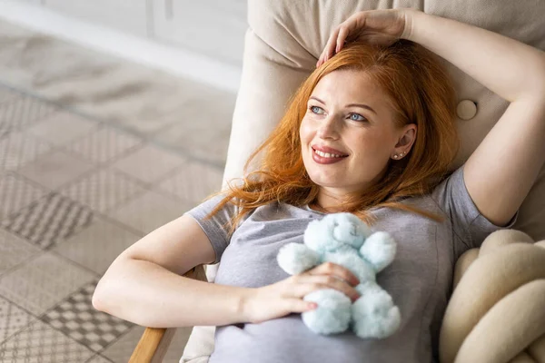 Top view happy young redhead pregnant woman lying on comfortable armchair with cute bear toy — 图库照片