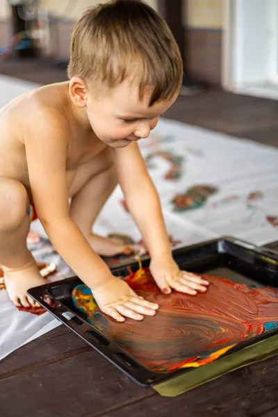 Niño dibuja con las manos y los pies con pinturas de dedos en una sábana grande en casa — Foto de Stock
