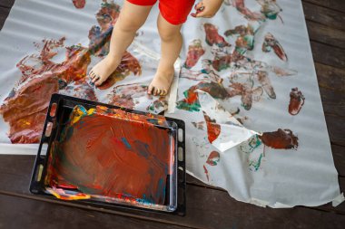 Küçük çocuk evde büyük bir çarşafa el ve ayaklarıyla parmak boyaları çiziyor.