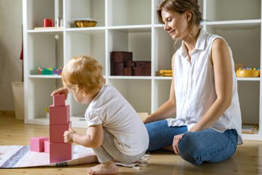 Young mother and little baby assembling pink cube Maria Montessori ecological materials clipart