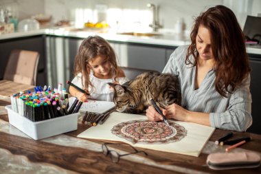 Genç, zayıf ve güzel bir anne güzel bir kızı ve Maine Rakunu kedisi mutfakta resim yapıyor. Dairesel desen mandala ve zen çizimi.