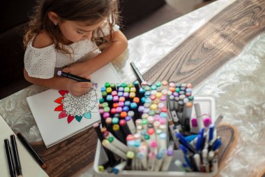 Küçük tatlı bir kız albümde çizim kalemleriyle dairesel bir mandala deseni çiziyor..