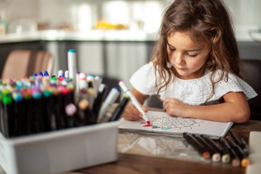 Küçük tatlı bir kız albümde çizim kalemleriyle dairesel bir mandala deseni çiziyor..