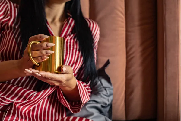Close-up beleza mãos femininas em pijama segurando caneca metálica dourada com bebida quente relaxante na cama — Fotografia de Stock