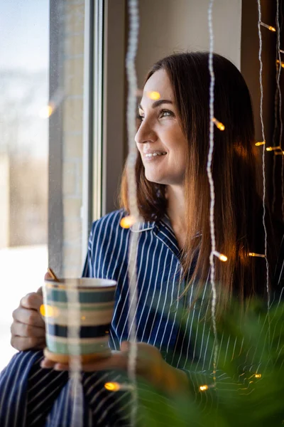 아침 커피를 들고 가랑을 든 채 집 창문 가까이에서 있는 잠옷을 입고 행복 한 가정 여성 — 스톡 사진