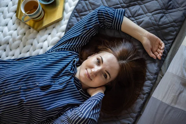 Top vista retrato de mulher doméstica relaxada feliz em pijama deitado na cama com chá e chocolate — Fotografia de Stock