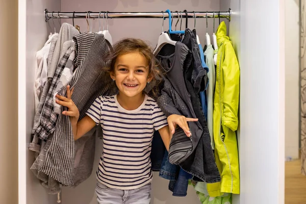 Портрет милая маленькая девочка позирует, прячась в шкафу с повешенной одеждой наслаждаясь счастливым детством — стоковое фото