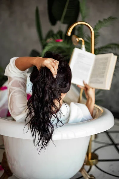 Atrás ver seductora mujer de moda leyendo libro de papel acostado en baño retro con pétalos de rosa spa —  Fotos de Stock