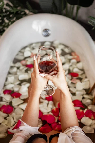 穿着黑色内衣的性感女人手捧着盛放在复古浴花花瓣中的高脚杯红酒 — 图库照片